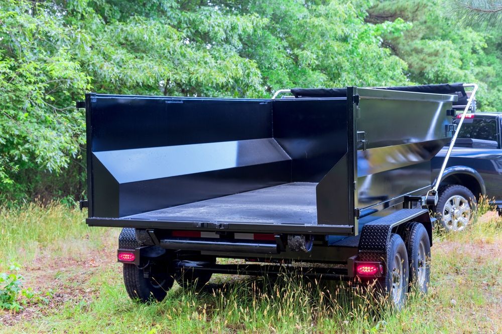 Black dump trailer with open bed in a grassy, wooded area.