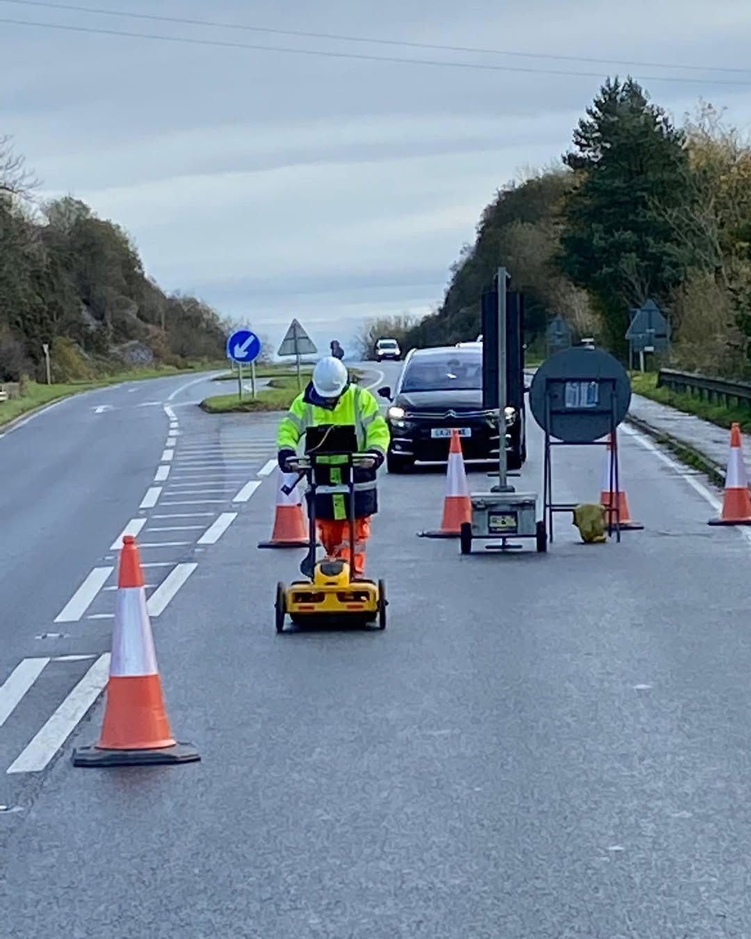 A worker in a safety vest uses a ground-penetrating radar on a road, flanked by cones and equipment.