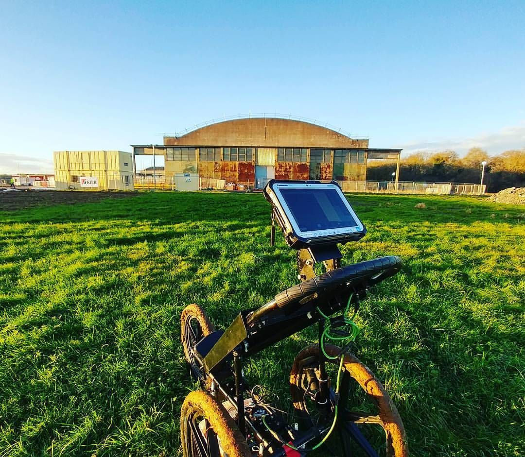 Ground-penetrating radar device on green field, old metal hangar in background, sunny day.