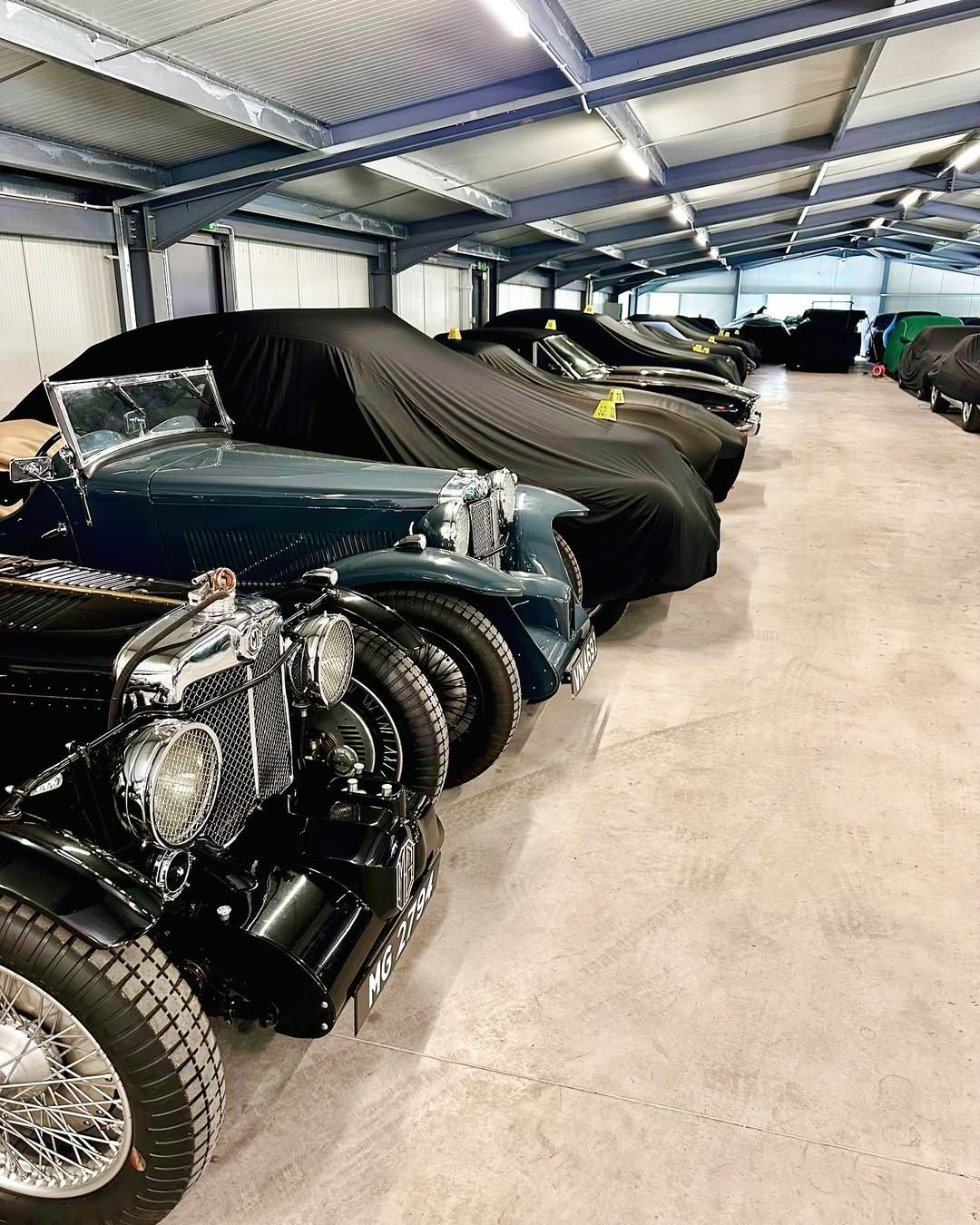Classic cars in a large, well-lit warehouse; several are covered with black covers.