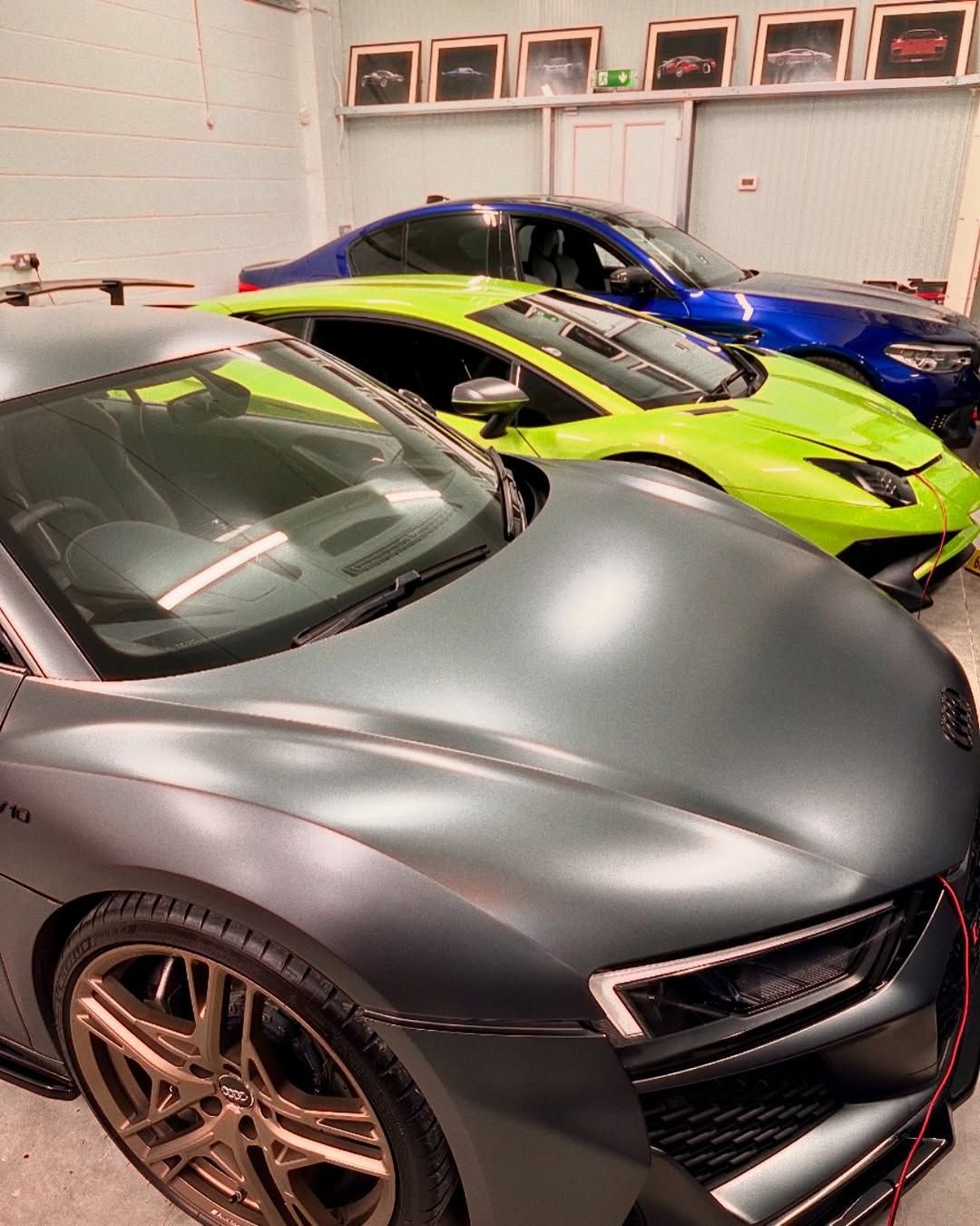 Gray Audi R8 sports car in a garage with a lime green Lamborghini and a blue car in the background.