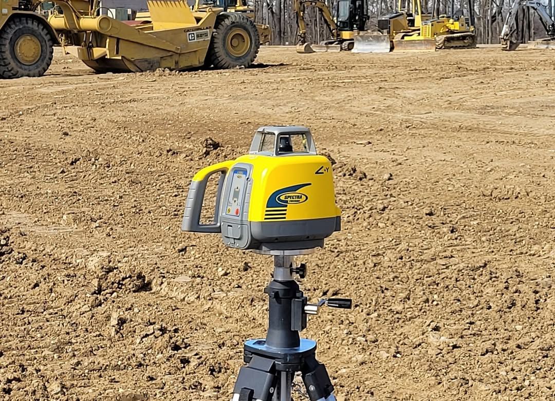 Yellow laser level on tripod at a construction site with heavy machinery in the background.