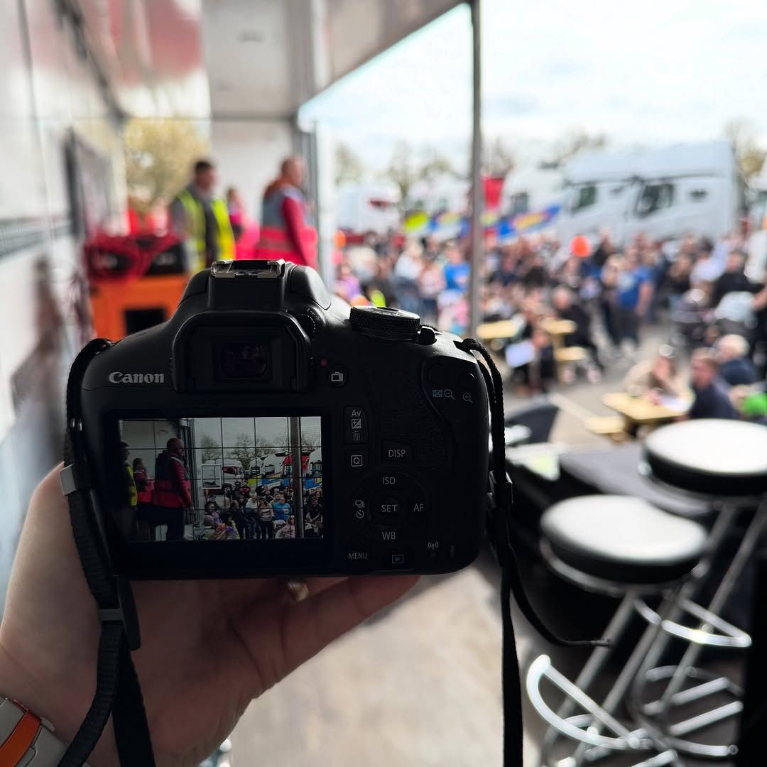 A person holds a Canon camera, the screen displaying a crowded event. Focus is on the event outside the camera.