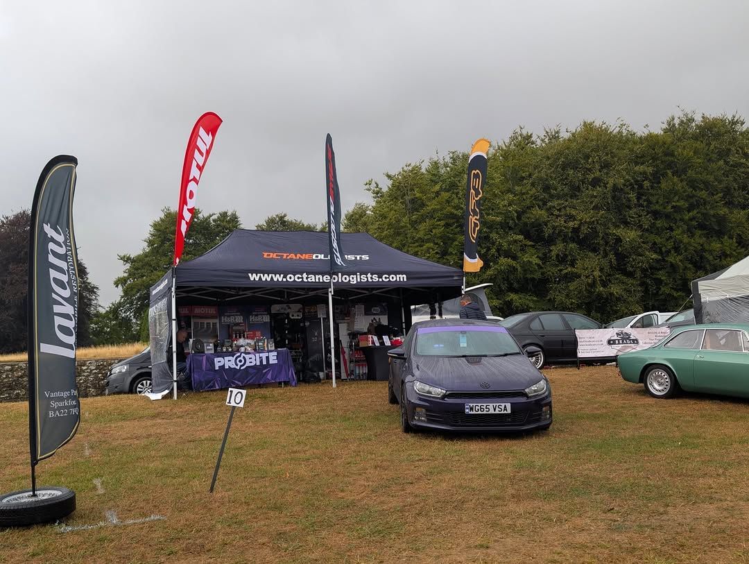 A car show tent with a dark blue car parked in front. Flags and merchandise are visible on a grassy field.
