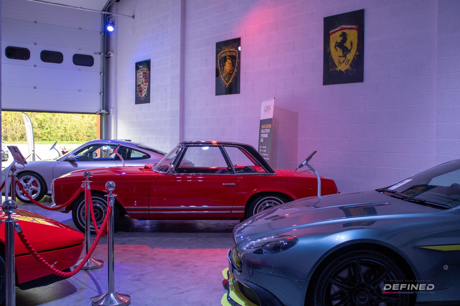 Garage with red classic car, blue Aston Martin, and Porsche. Ferrari and Lamborghini posters on the wall.