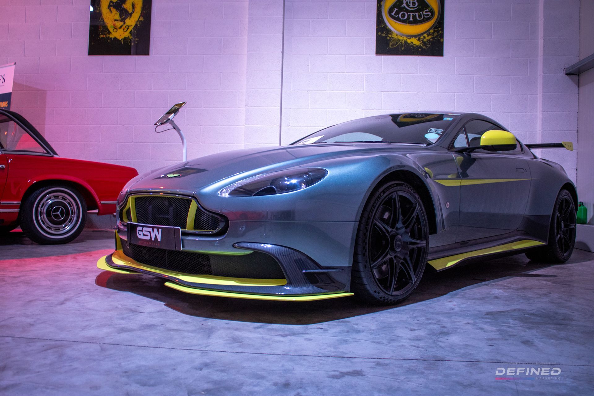 Gray Aston Martin sports car with yellow accents, parked in a garage with a red car visible in the background.
