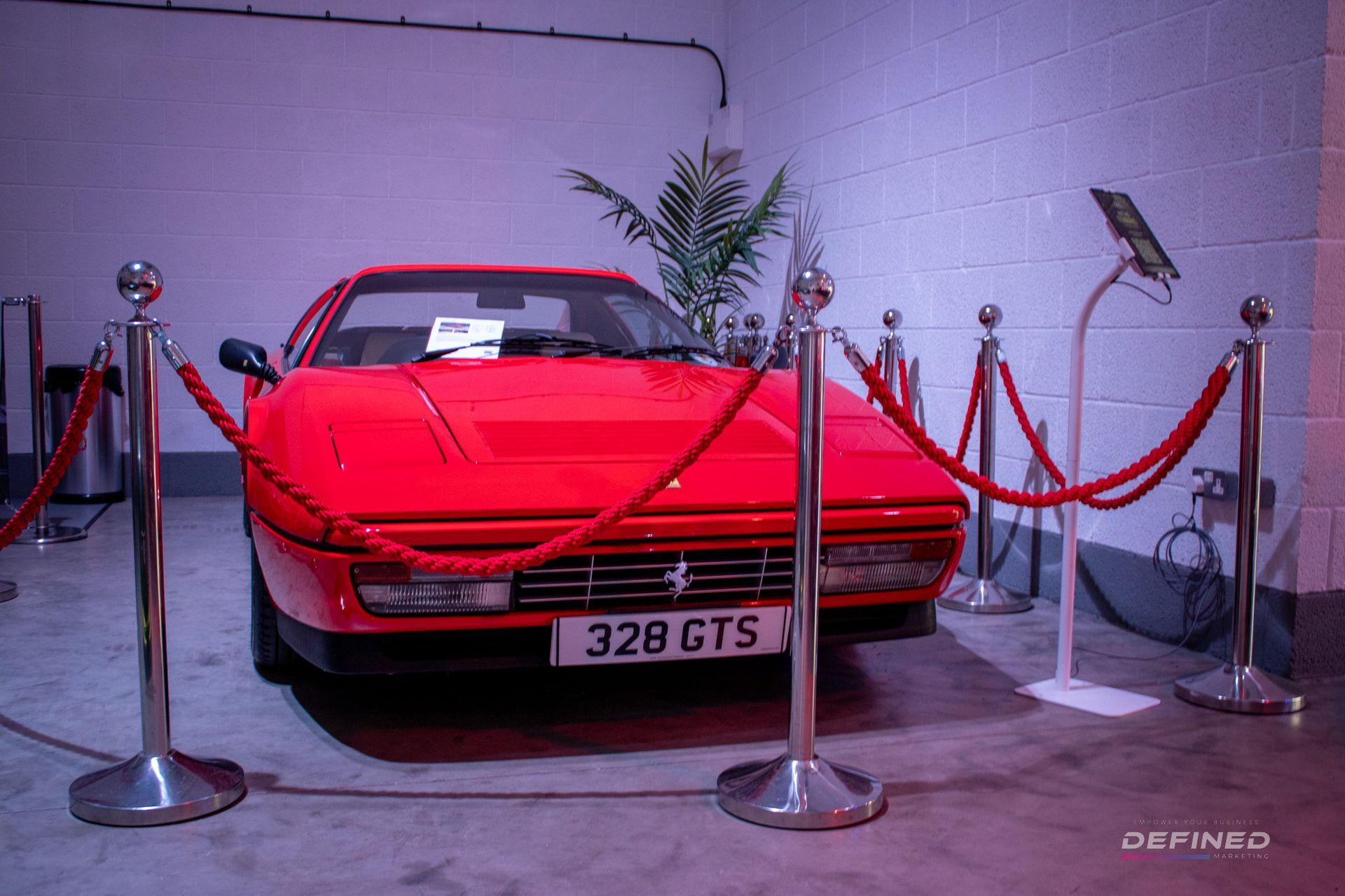 Red Ferrari 328 GTS behind velvet ropes in a showroom.