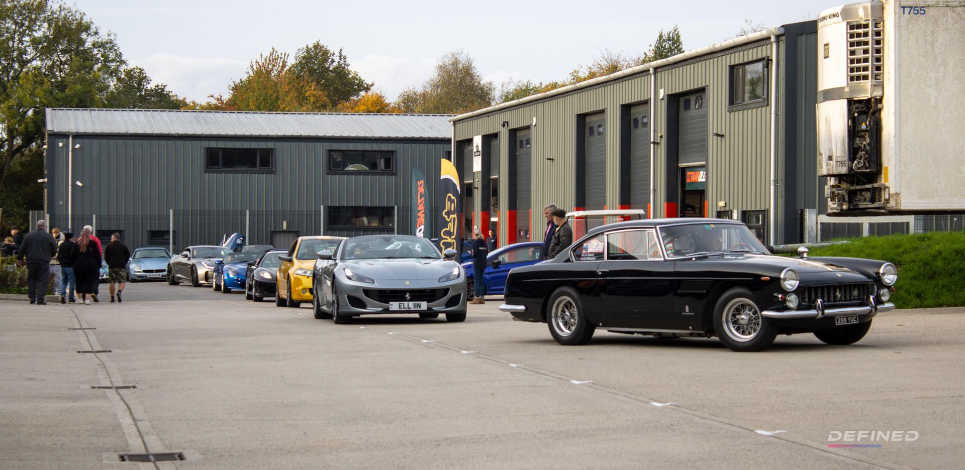 A black vintage car leads a line of modern sports cars, parked near industrial buildings.