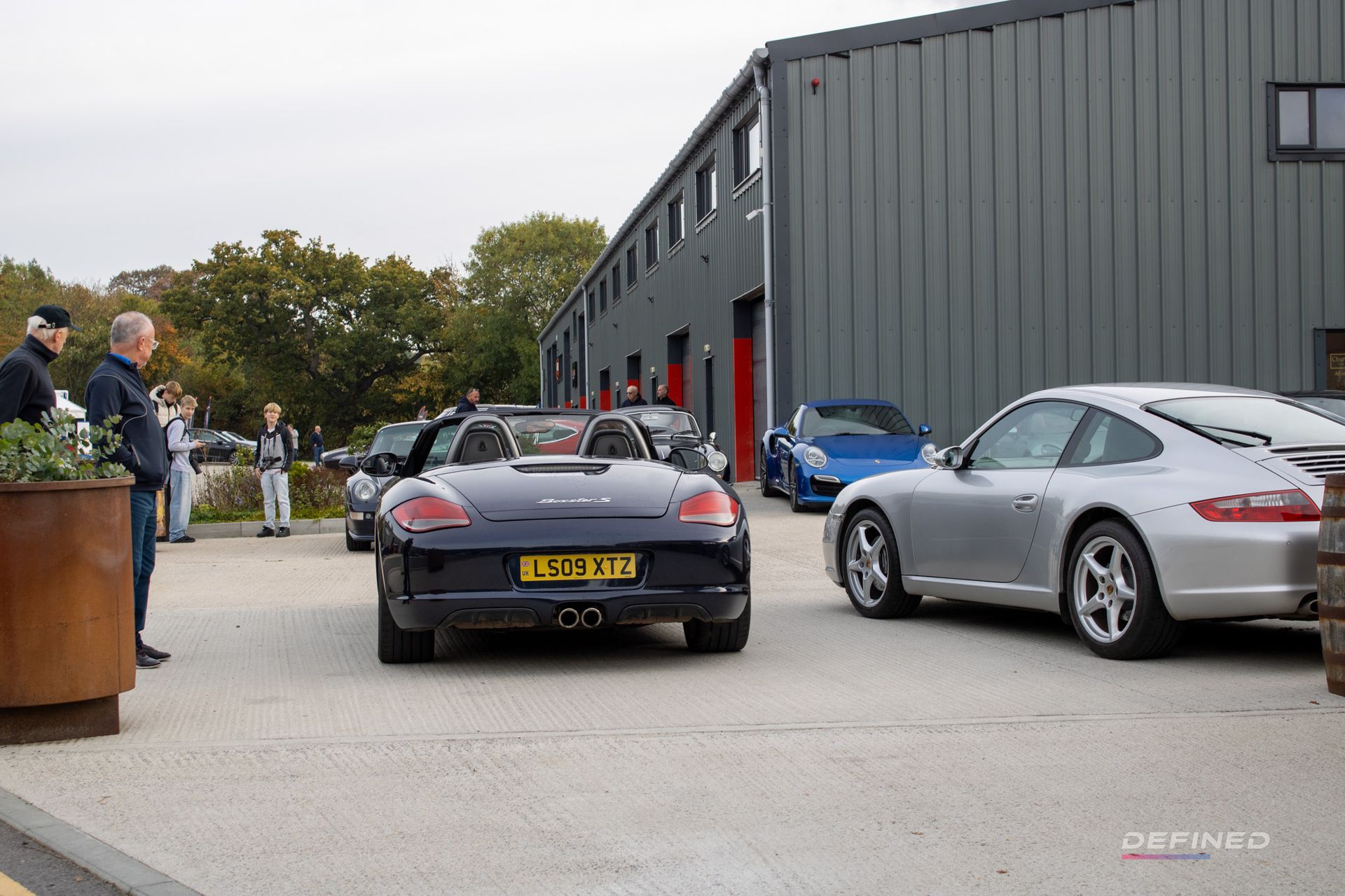 A line of luxury cars, including a blue Porsche Boxster, in front of a building. People stand nearby.