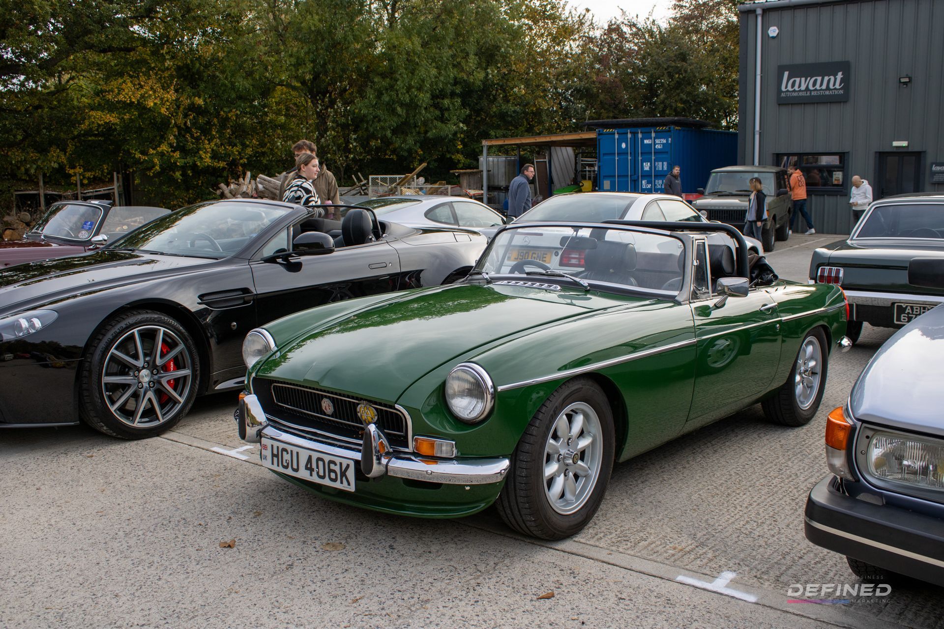 Green MG B roadster, black Aston Martin, and other cars parked outside a building.
