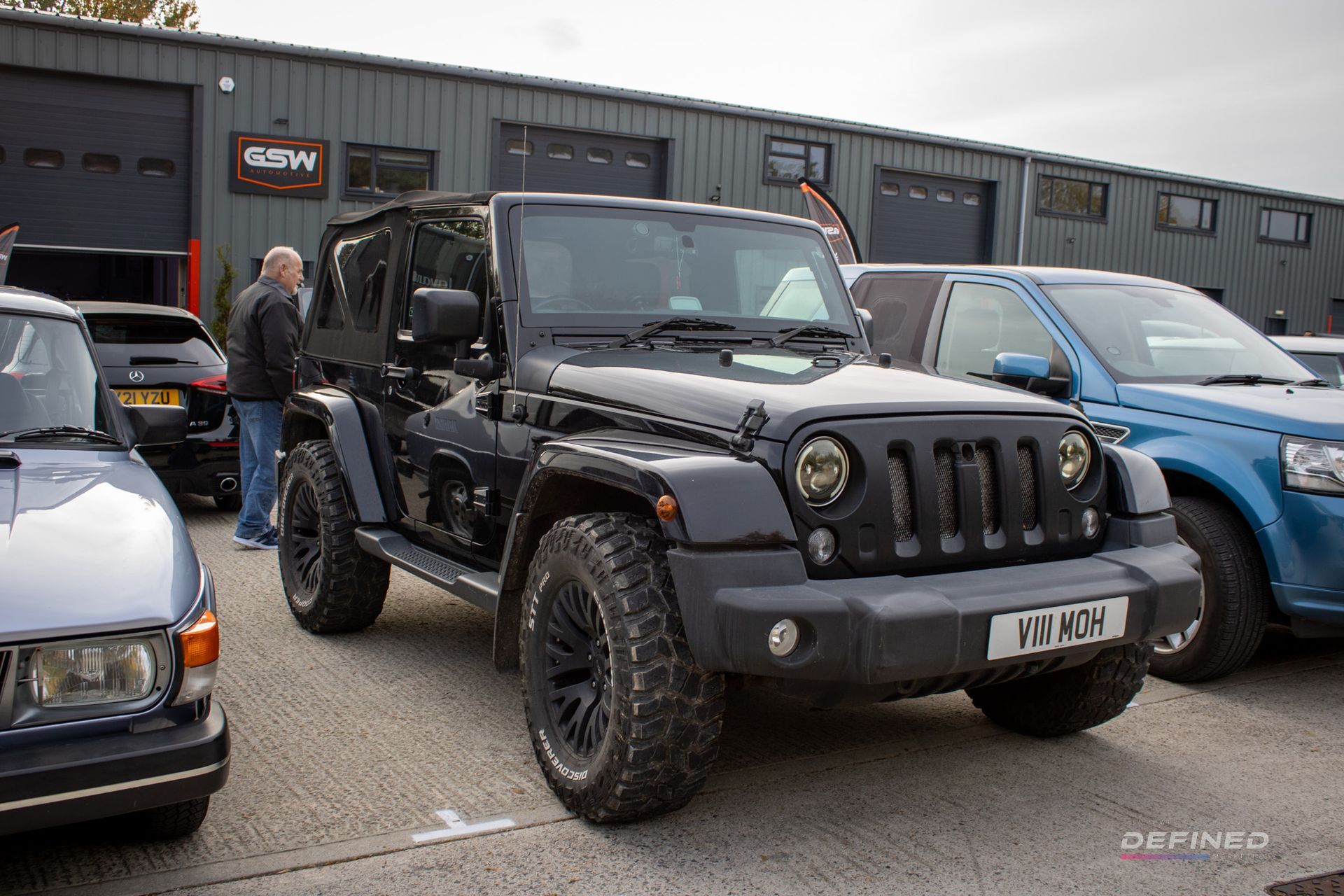 Black Jeep Wrangler parked in front of a building; other cars and a person are visible in the background.