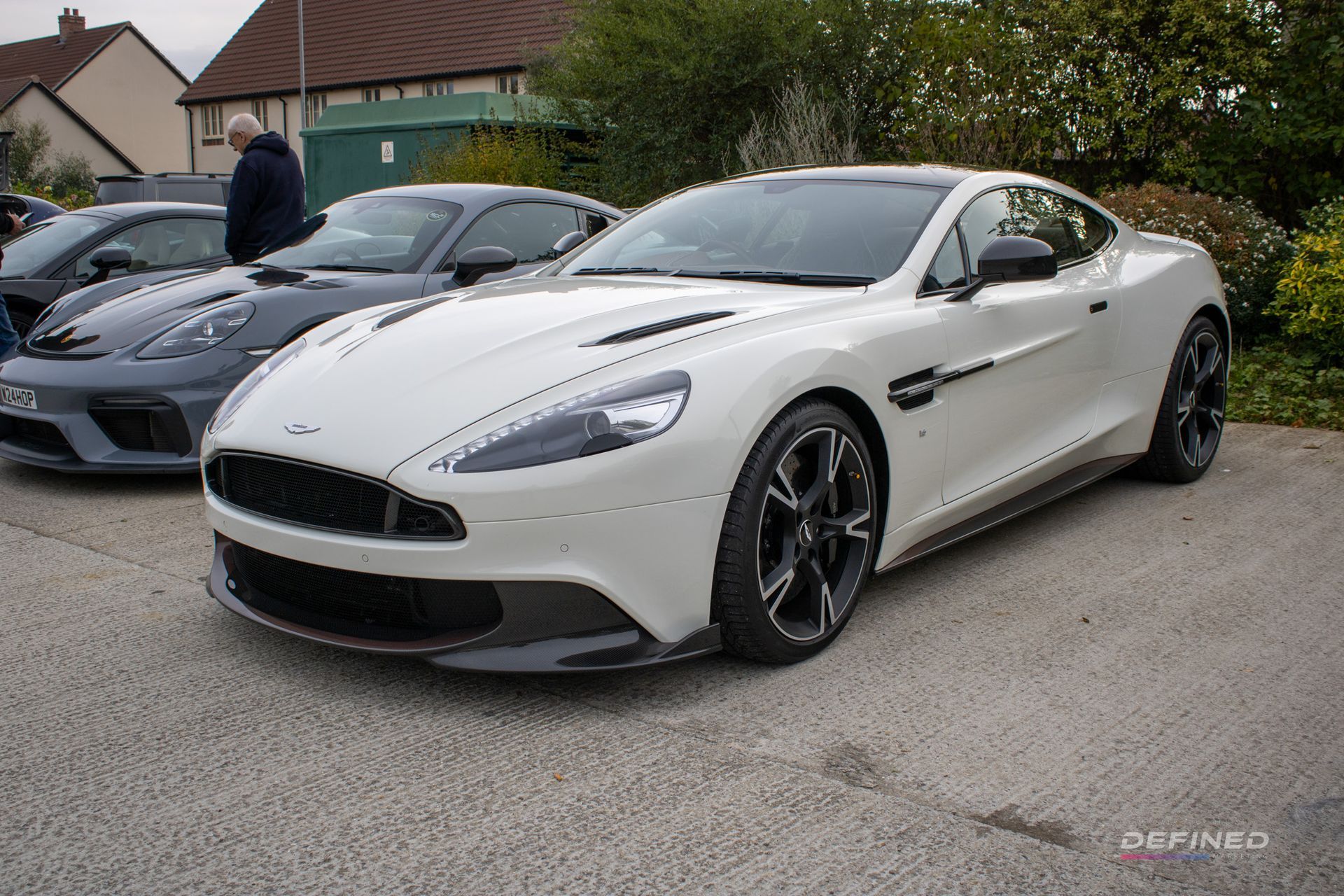 White Aston Martin sports car parked in a lot, other cars and people visible.