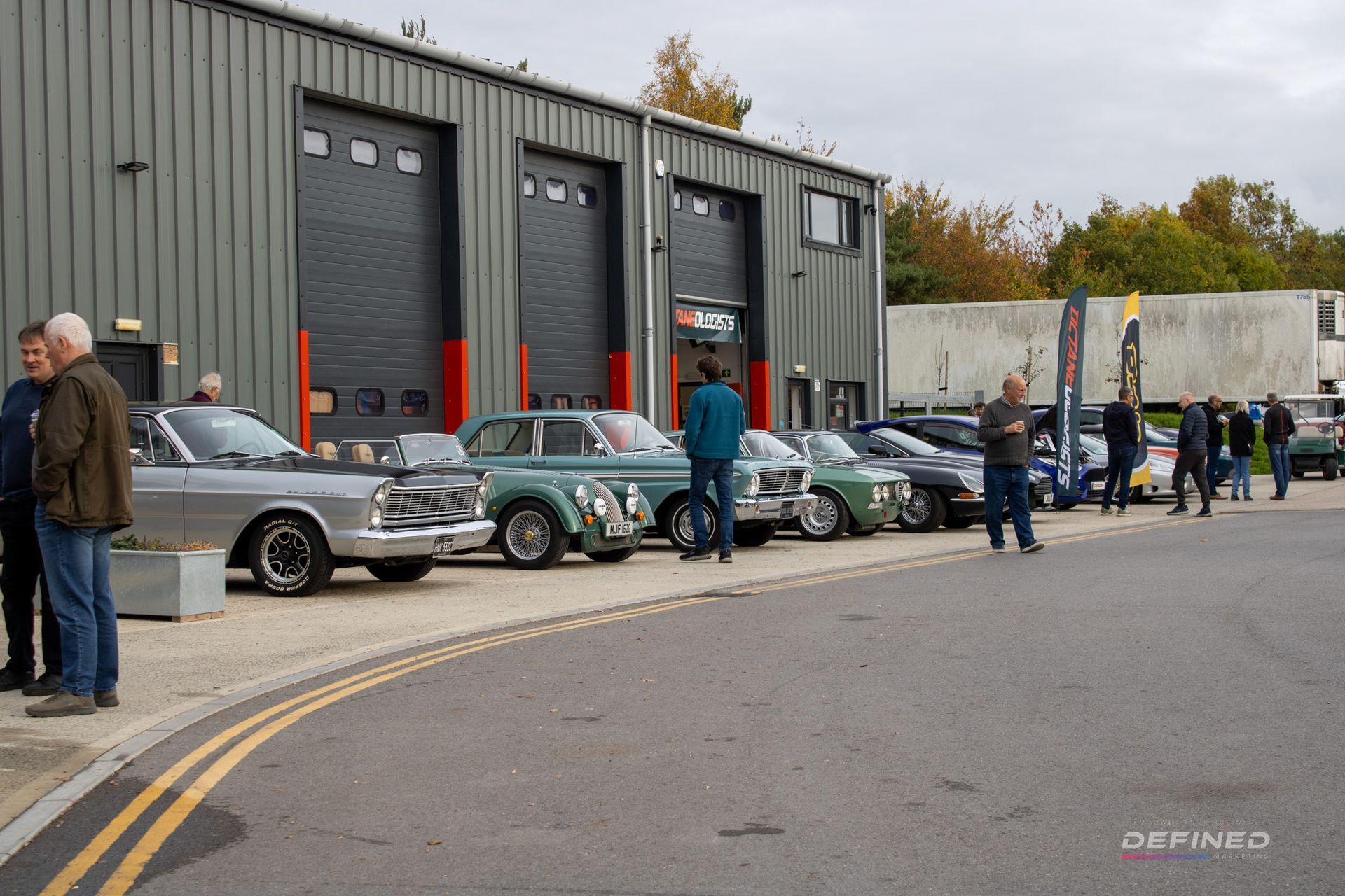 Classic cars parked outside a gray industrial building; people standing around.