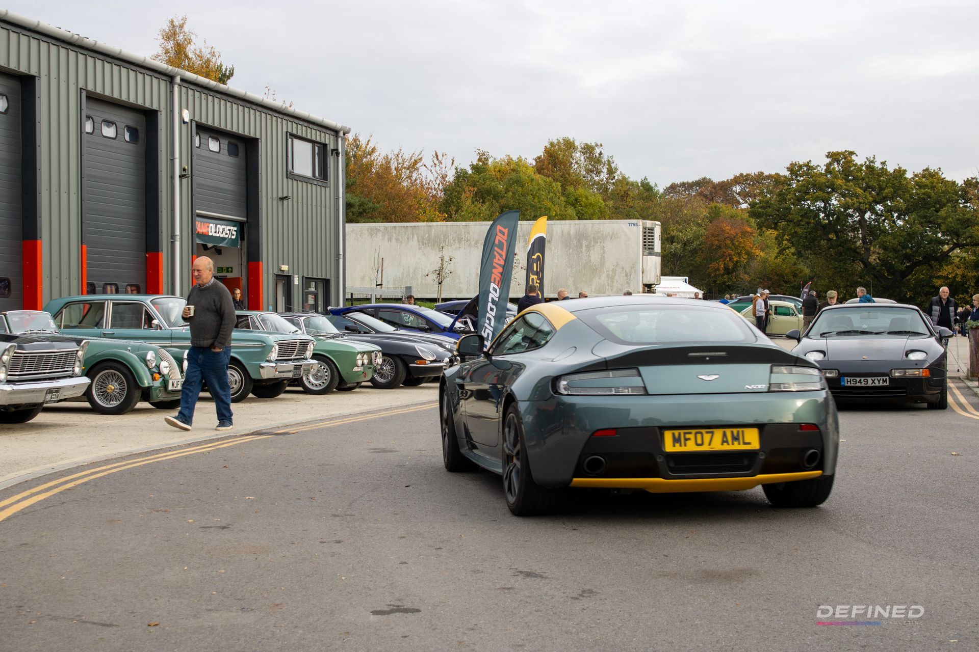 An Aston Martin sports car in front of a garage, surrounded by other classic cars. Man walks by.