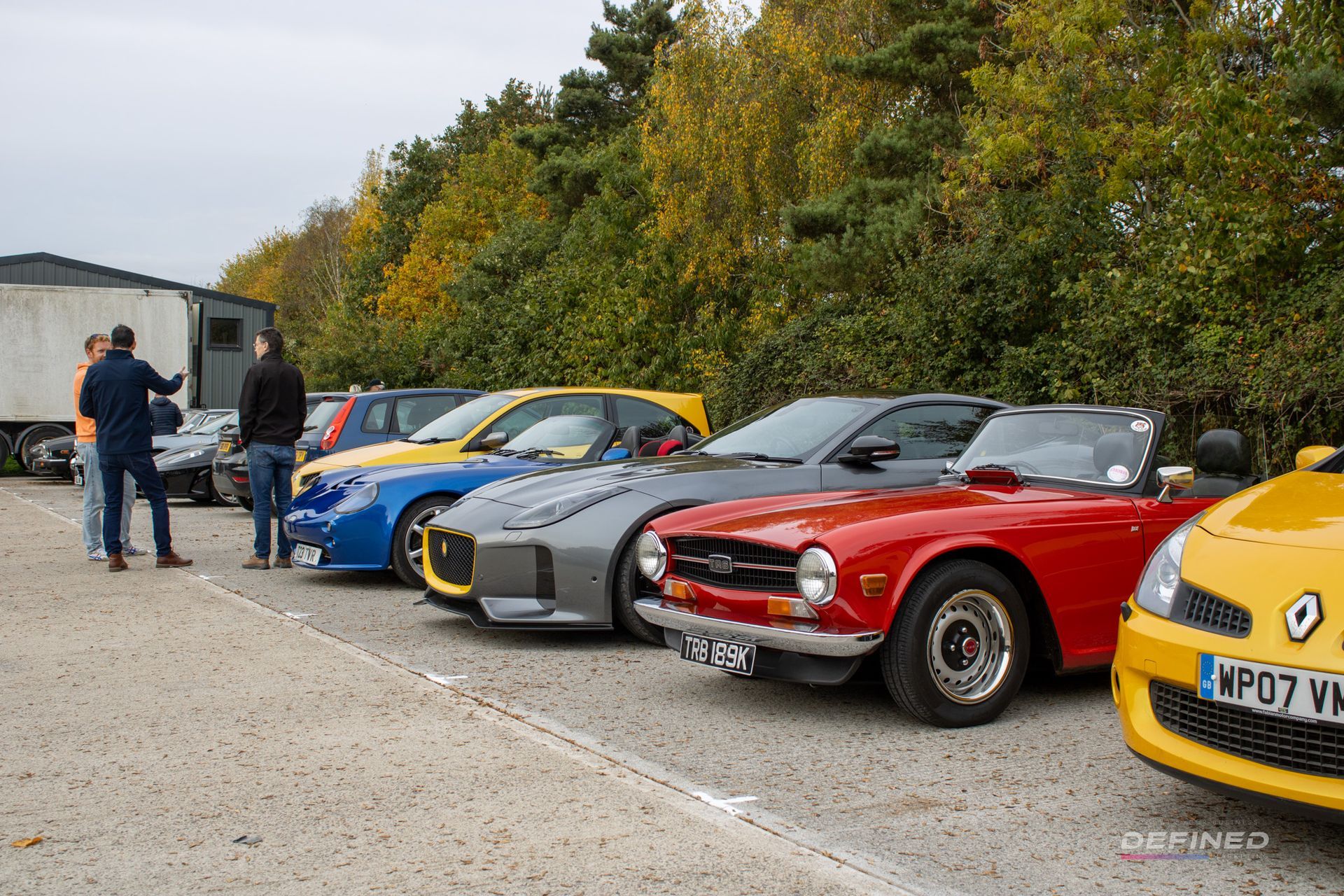 Cars parked in a row, including a red convertible and yellow hatchback, in a lot with people nearby.