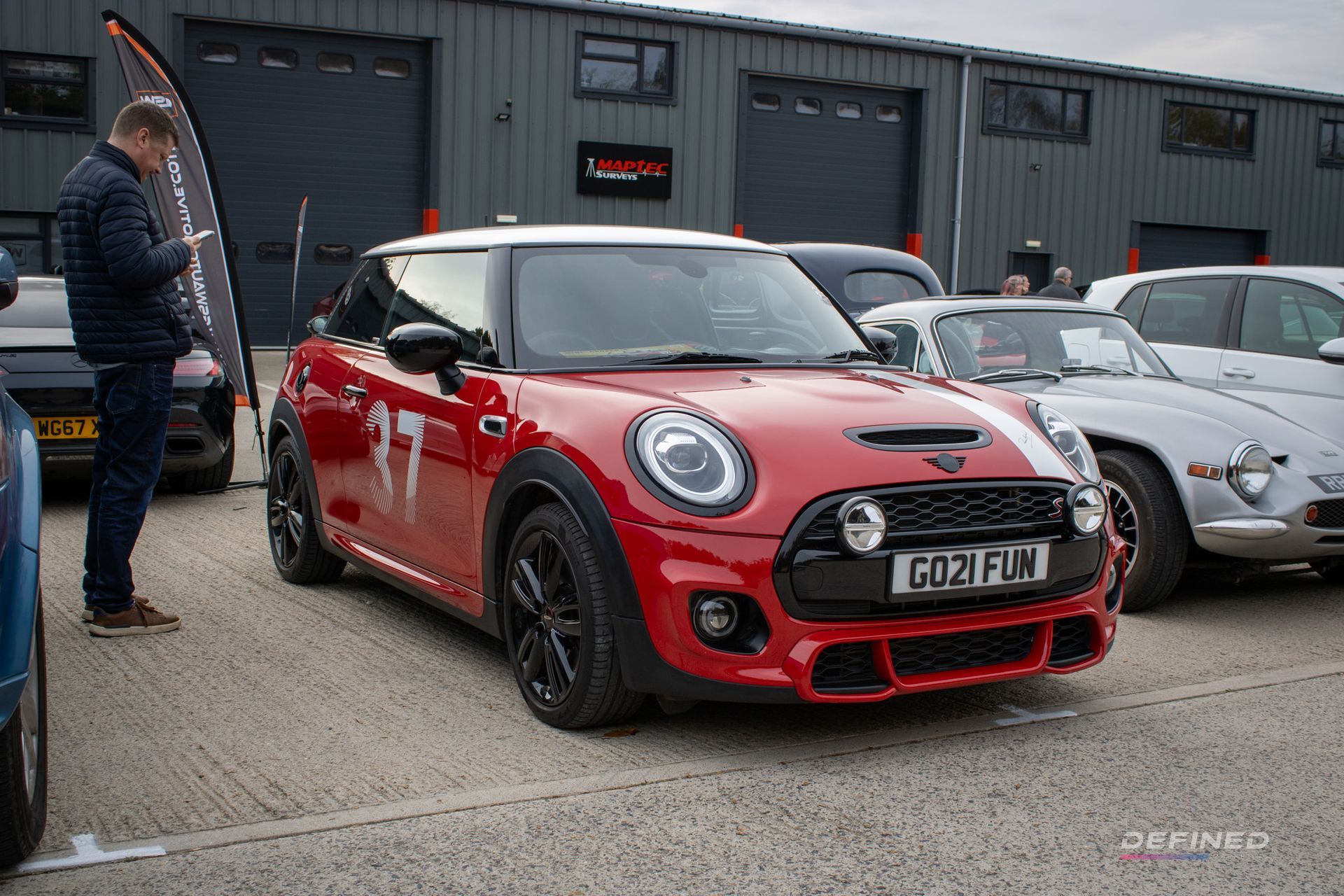 Red Mini Cooper with black accents parked outdoors, person looking at their phone nearby.