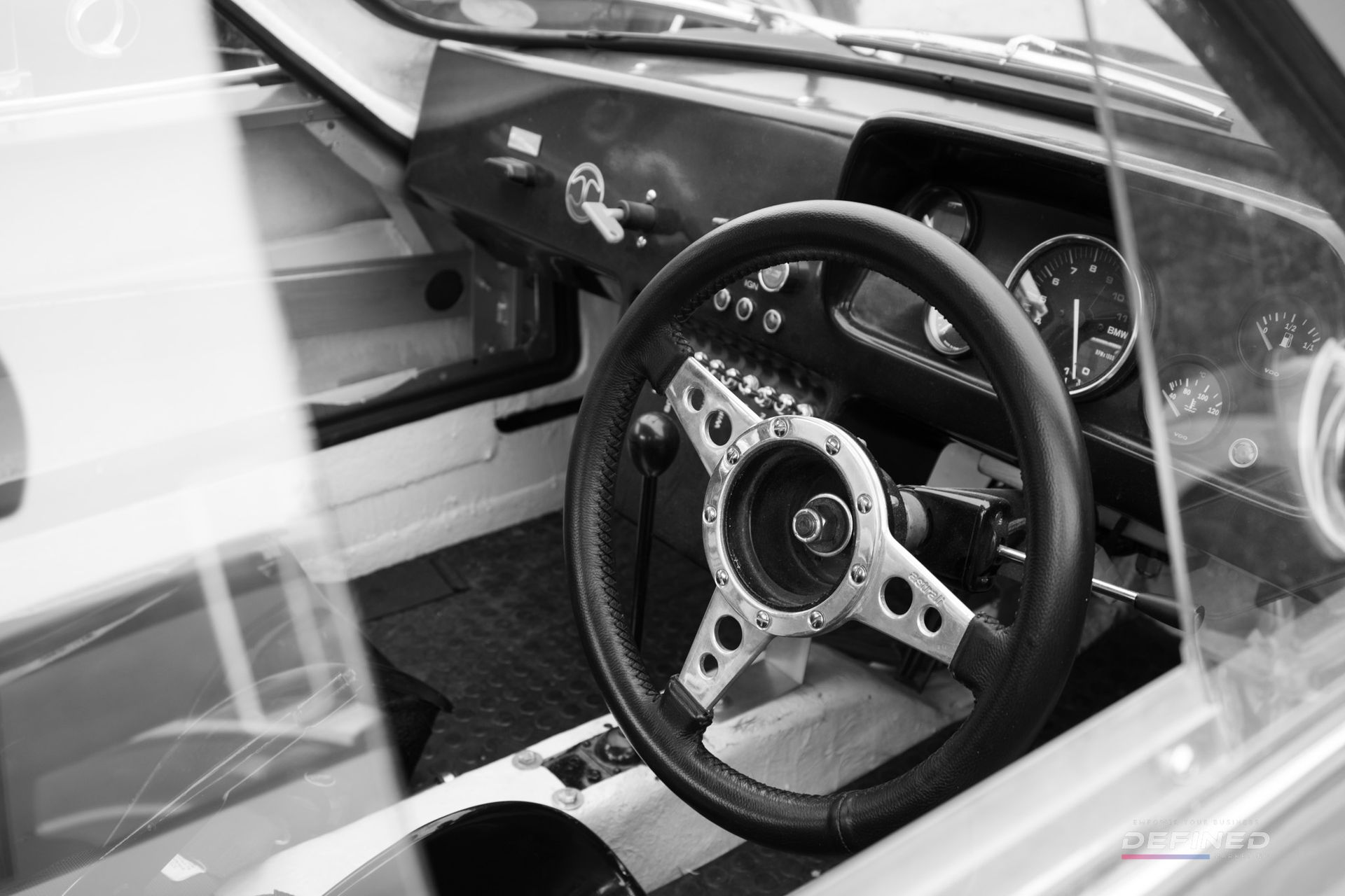 Black and white interior shot of a classic car, focusing on the steering wheel, dashboard, and windshield.
