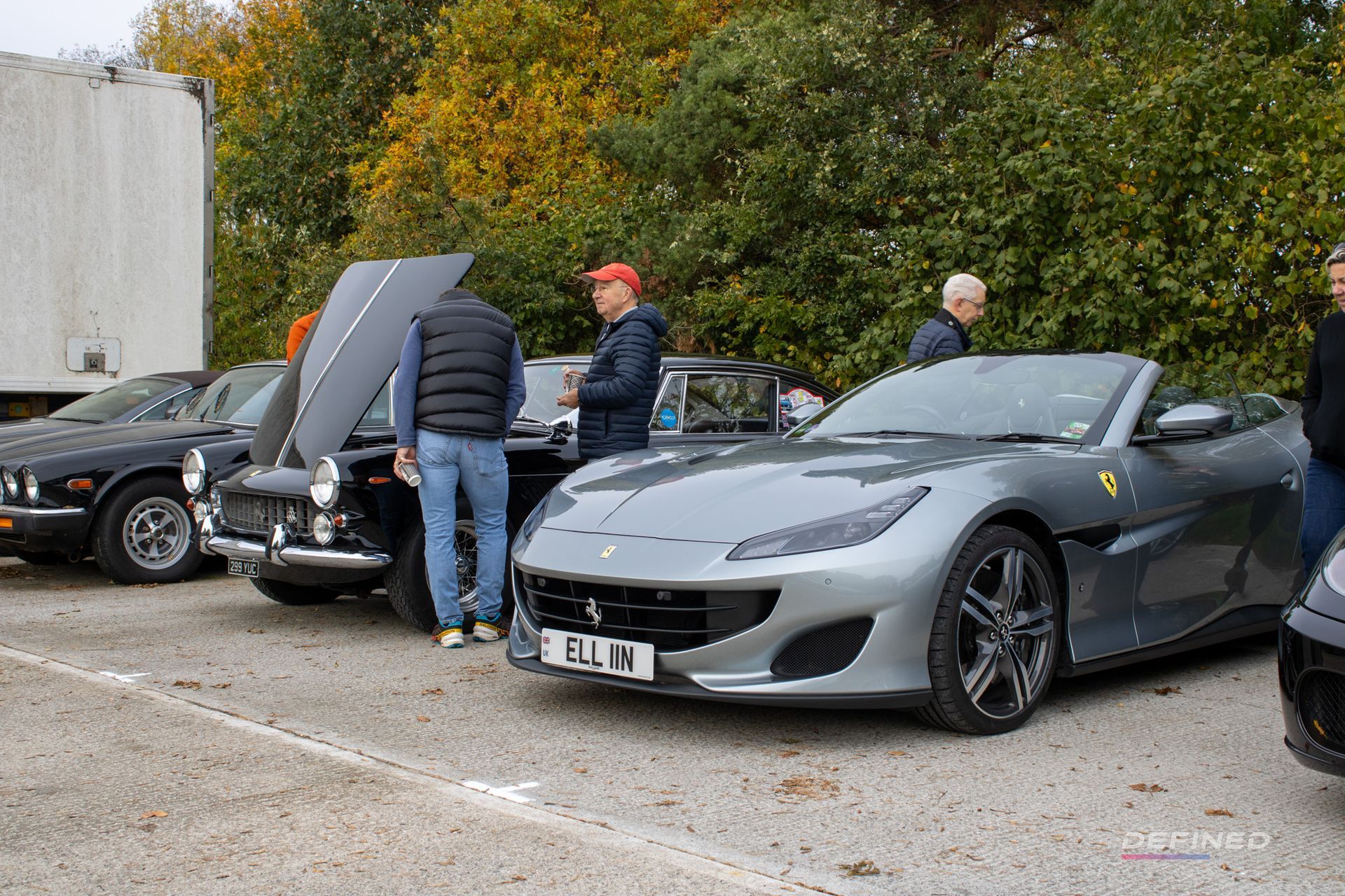 Several cars parked, including a silver Ferrari. People looking at the cars in an outdoor setting.