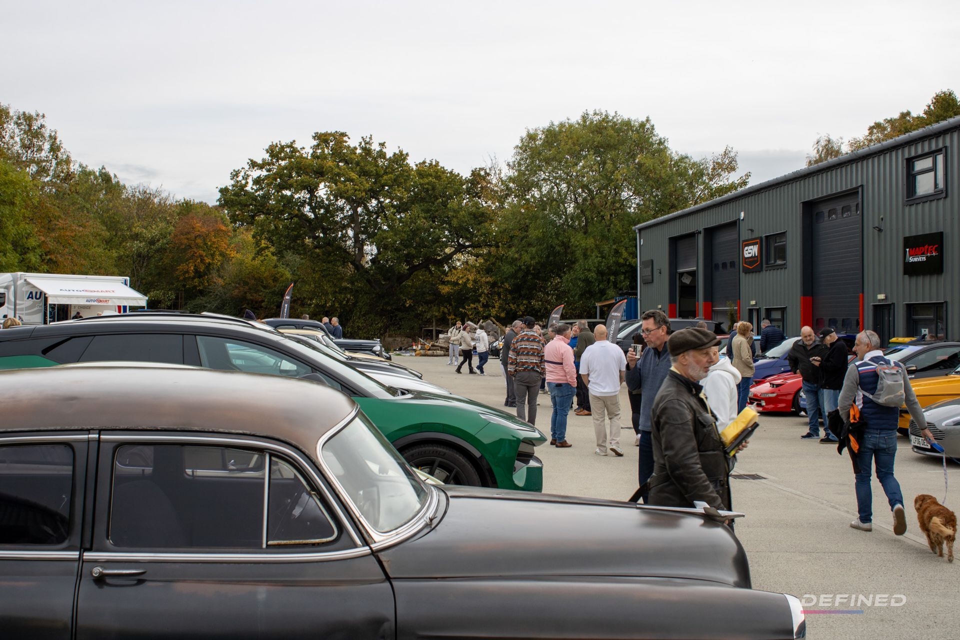 Car show with various vehicles and people gathering in front of a gray building on an overcast day.
