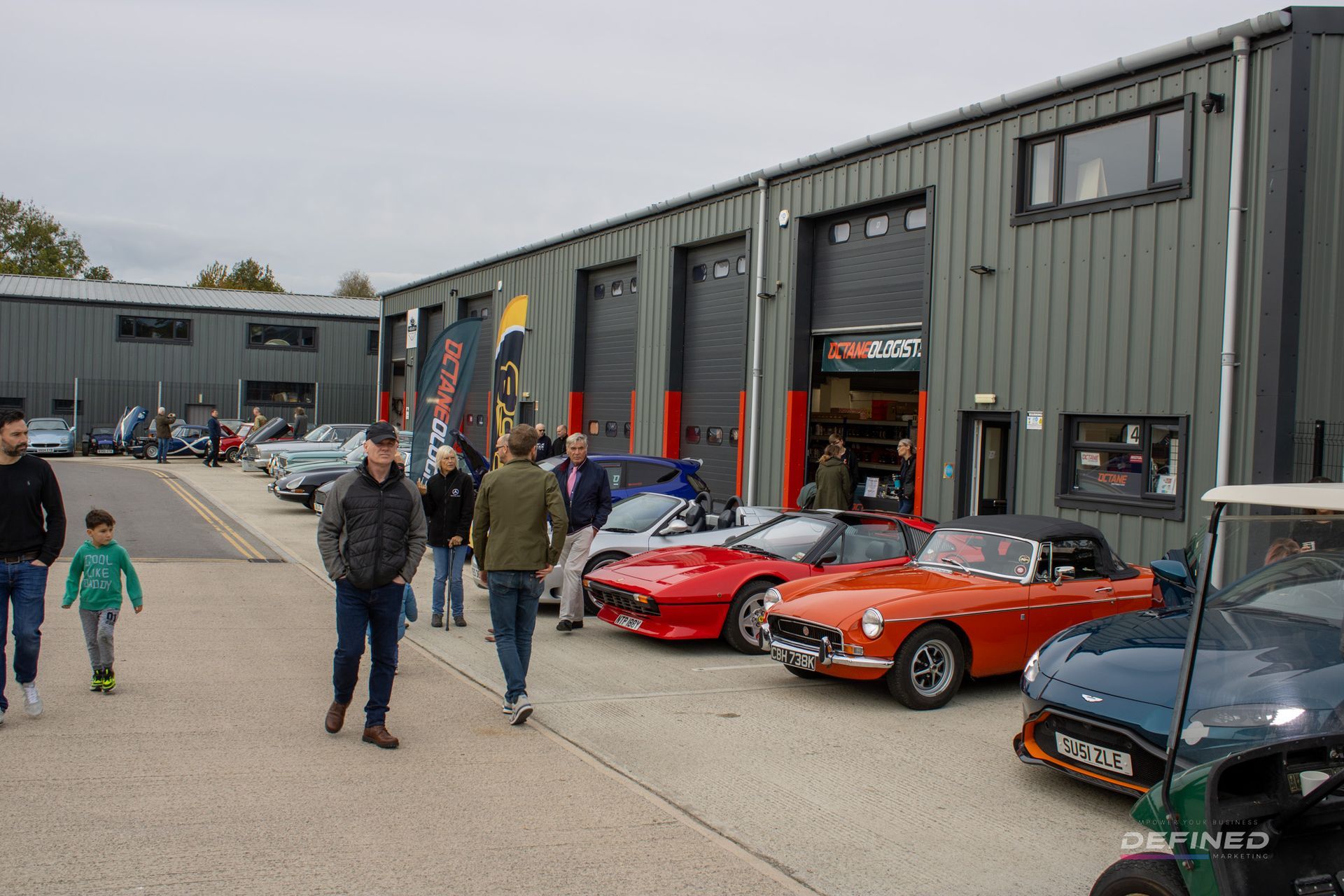 People viewing classic cars parked outside a gray industrial building on a cloudy day.