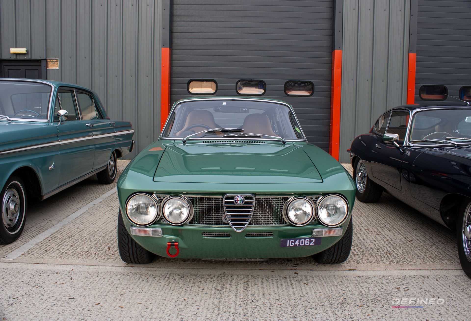 Green vintage Alfa Romeo car parked in front of a gray garage door, with other classic cars on each side.