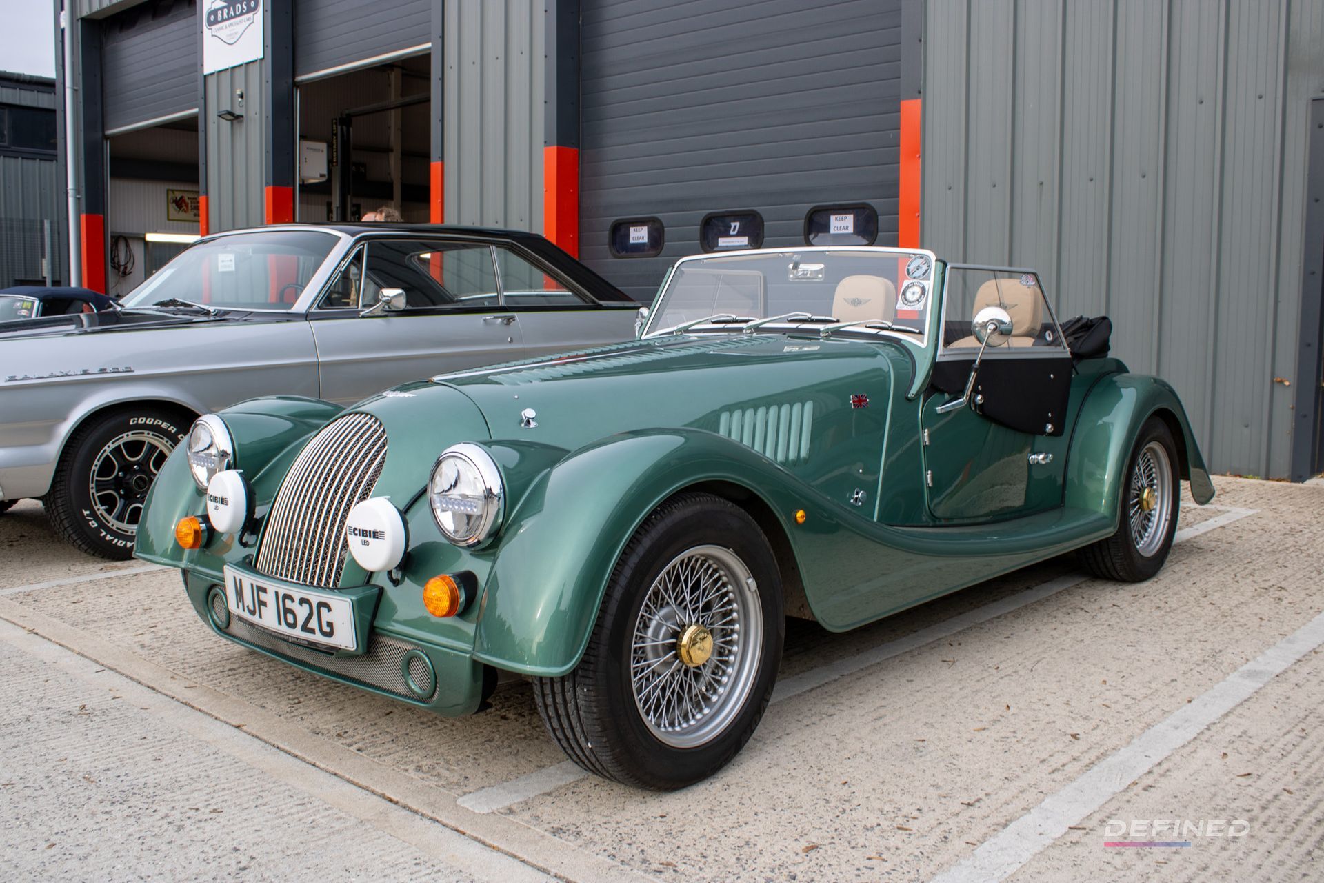 Green Morgan sports car parked in front of a garage, with another car visible behind.