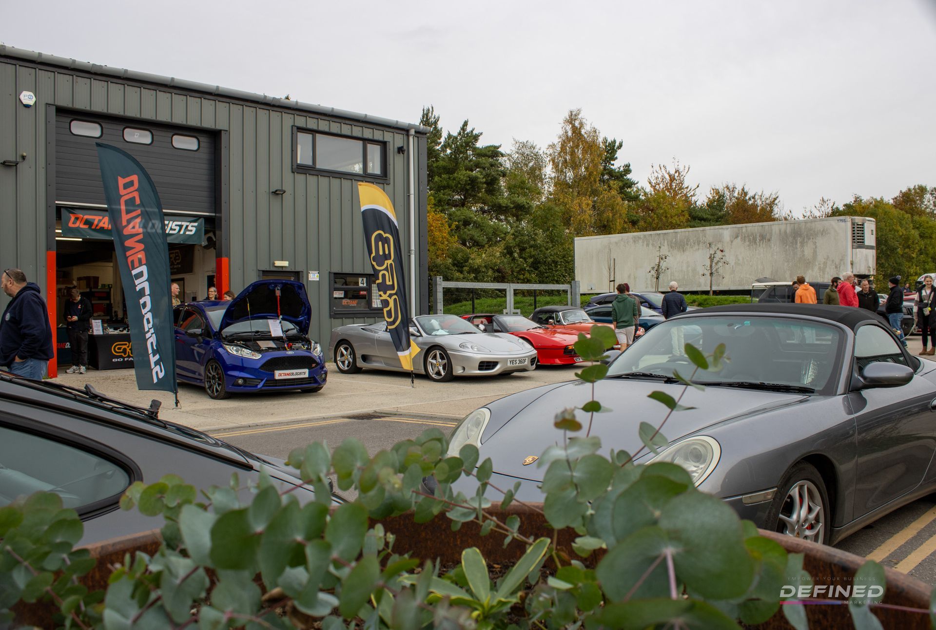 Cars parked outside a garage, car show. People gather. Gray and blue cars prominent. Cloudy day.