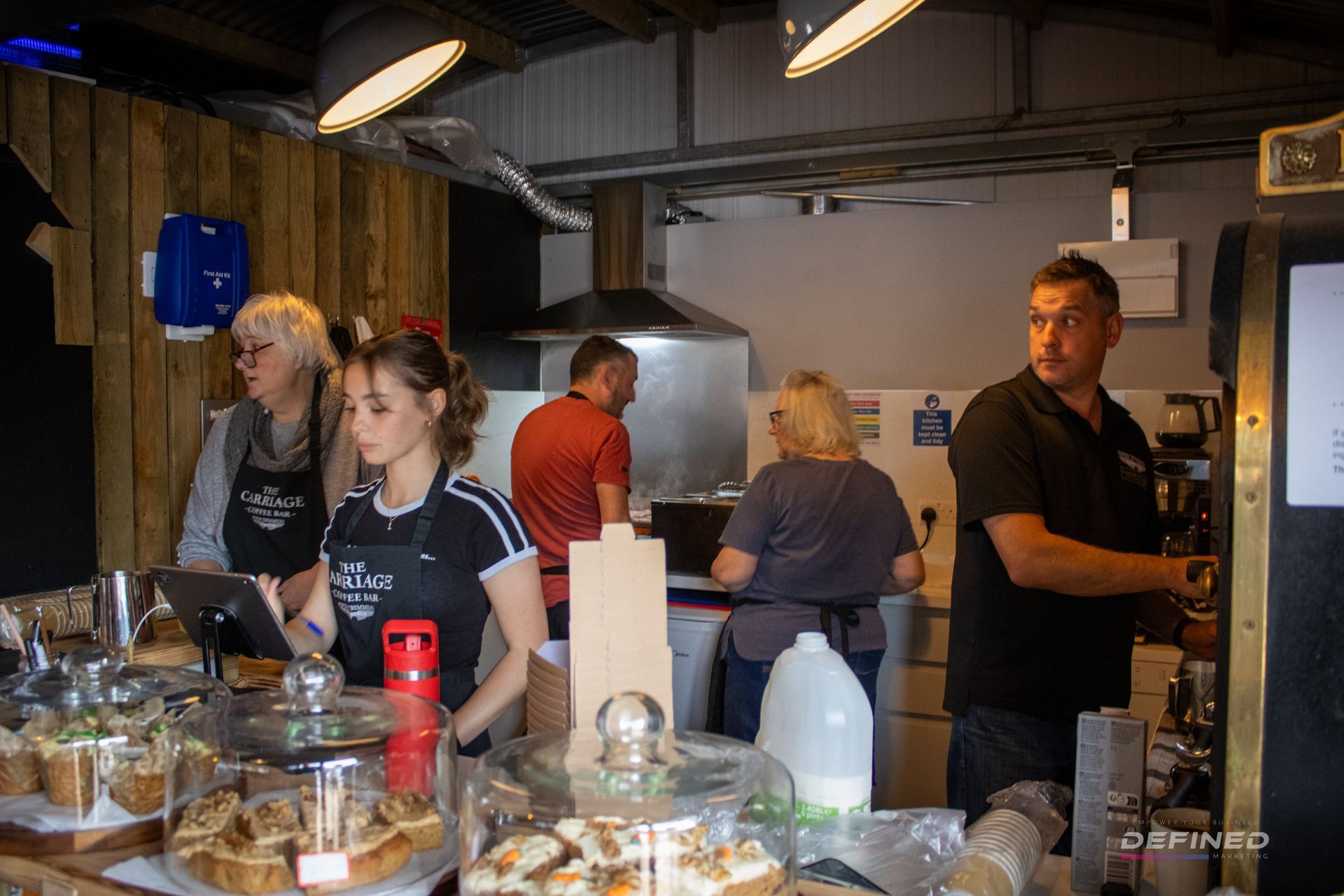 People working behind a cafe counter: two taking orders, others preparing food, stainless steel vent hood.