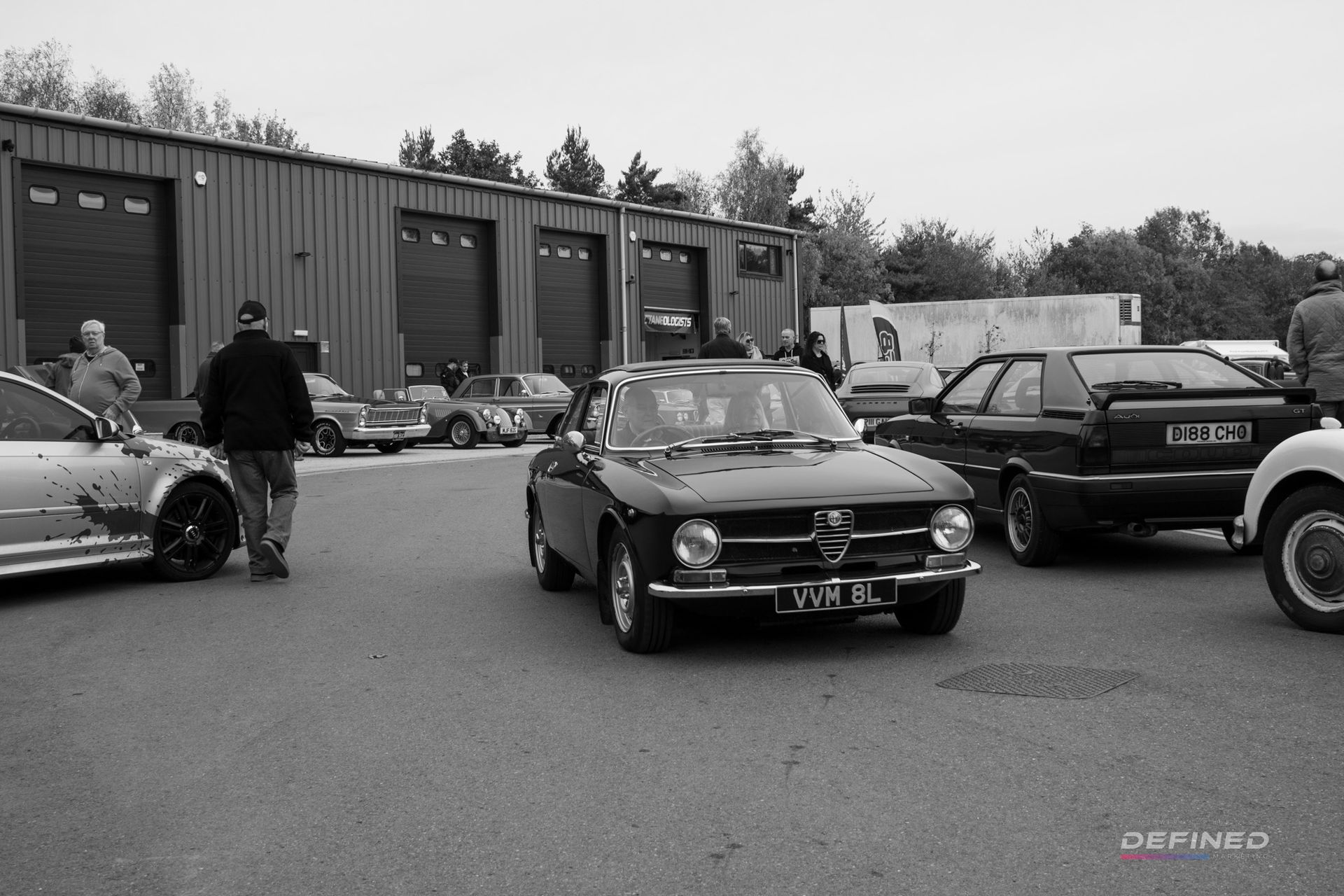 Black and white photo of vintage cars parked in front of a building; people standing around.