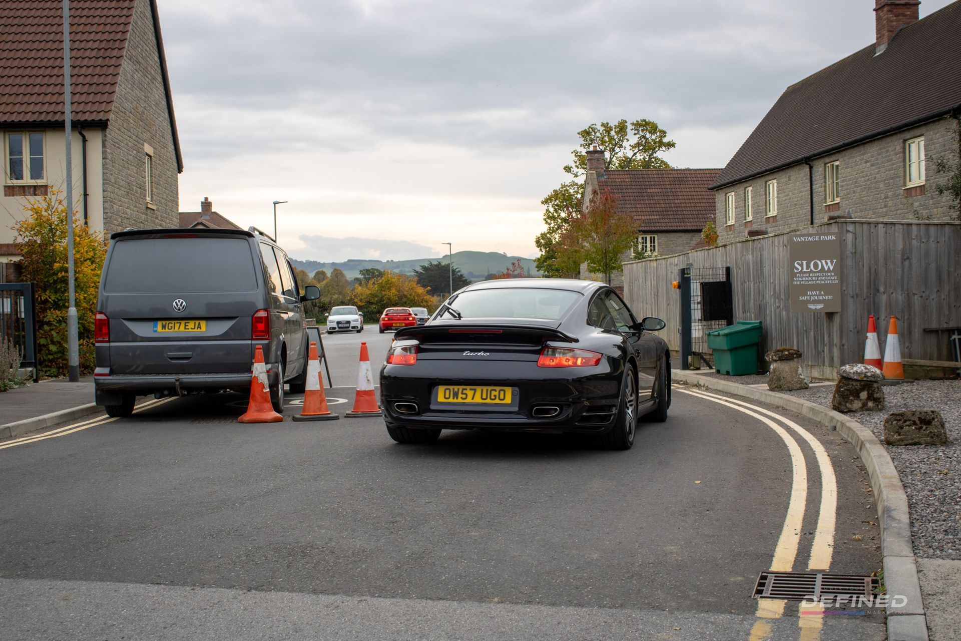 Black sports car on a curved road, orange cones separate it from a van. Buildings line the road, overcast sky.