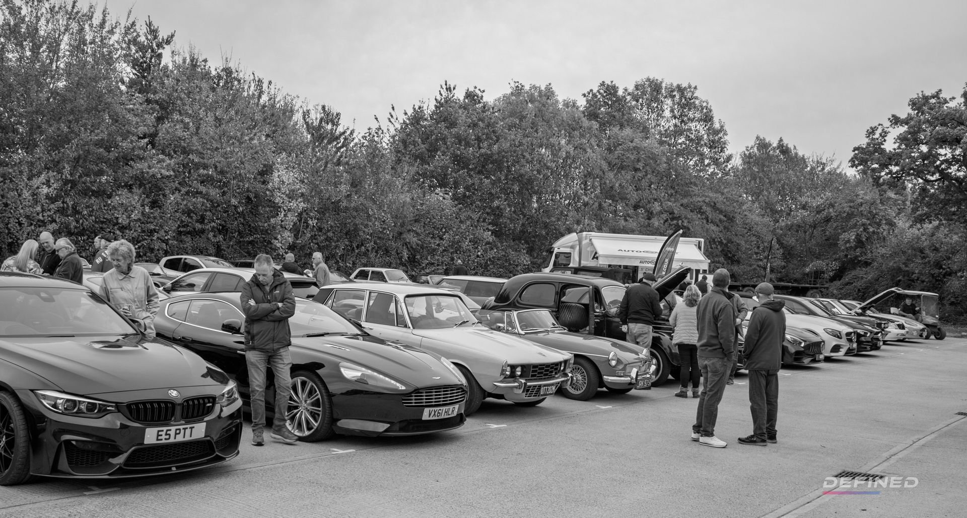 Black and white photo of a car meet. Several cars parked in a row with people gathered around.