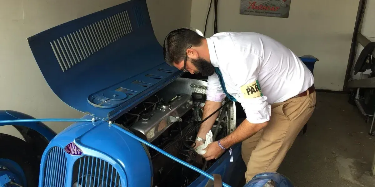 Man in white shirt and khaki pants working on a blue vintage car's engine, in a garage.
