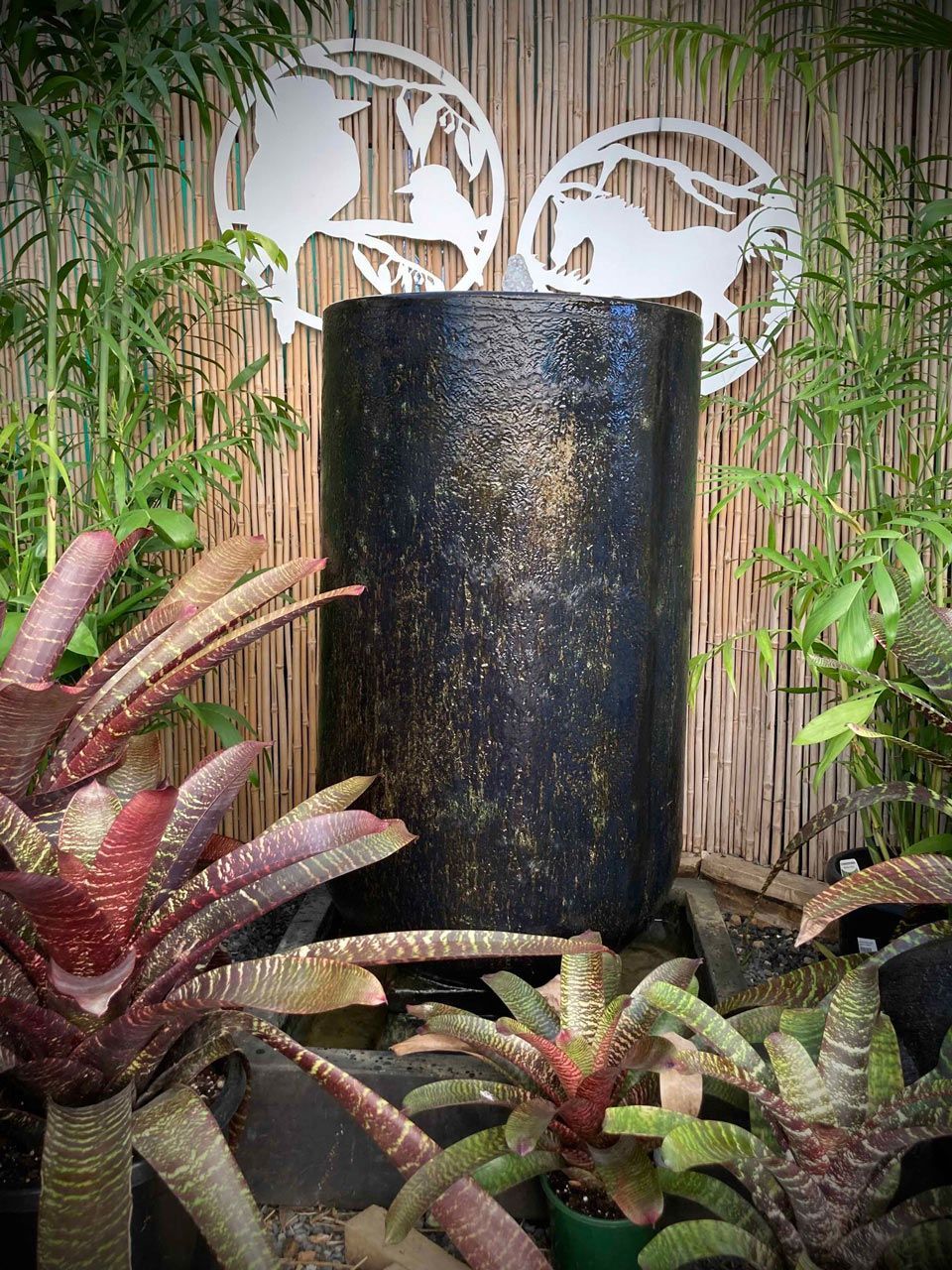A water fountain is surrounded by plants and a bamboo fence.