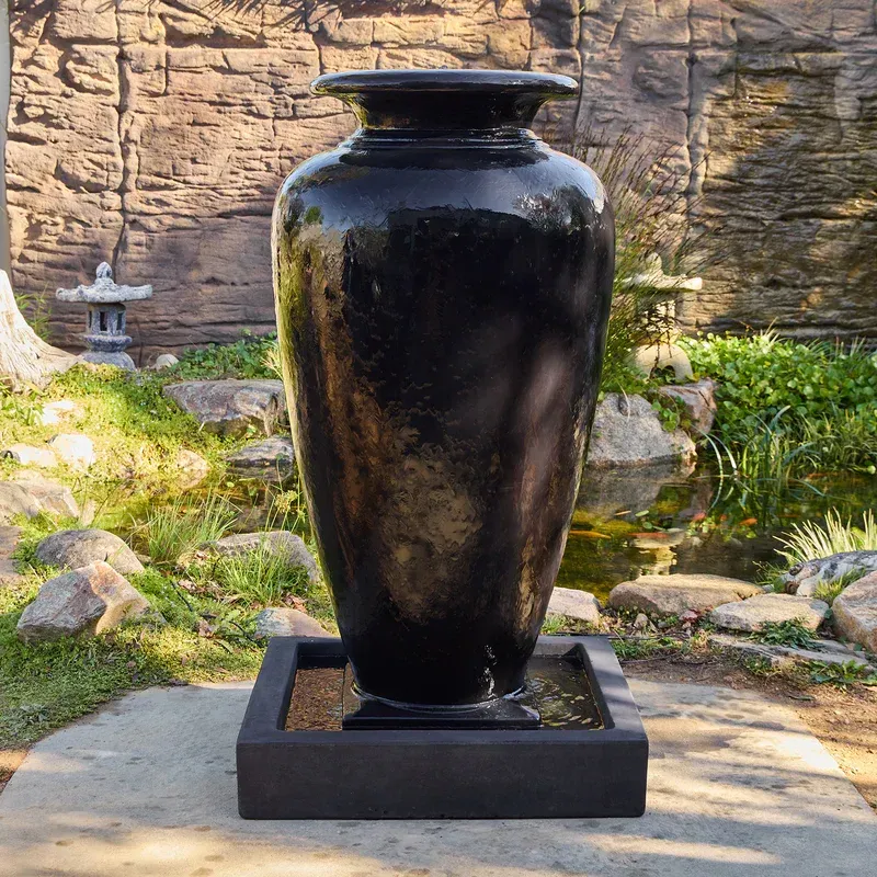 A large black vase is surrounded by plants in a garden.