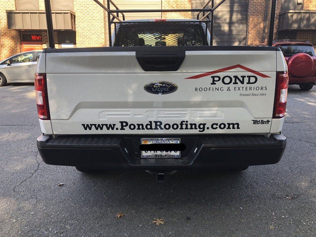 A white truck with a roofing company sticker on the back is parked in a parking lot.