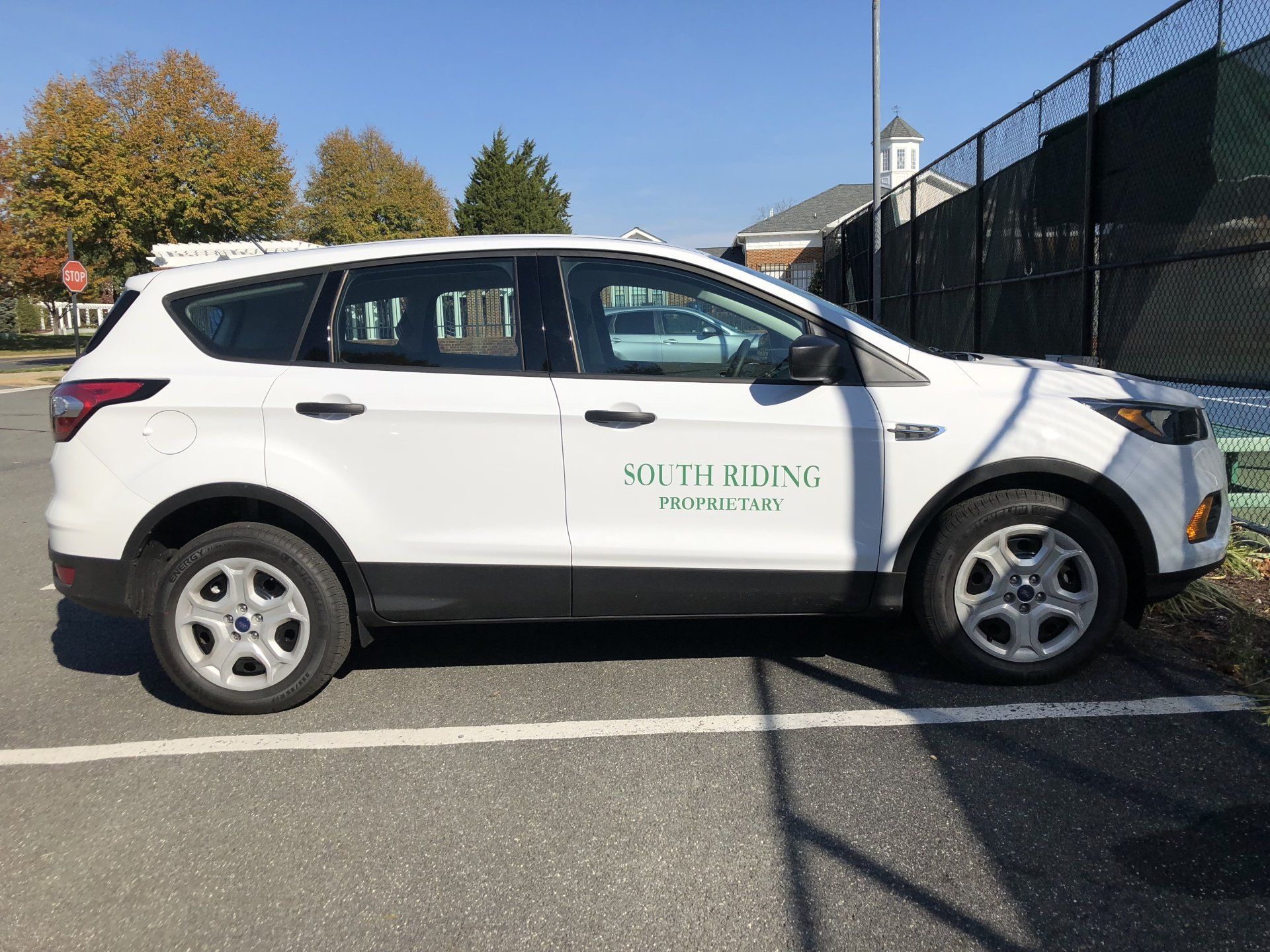 A white suv is parked in a parking lot next to a tennis court.