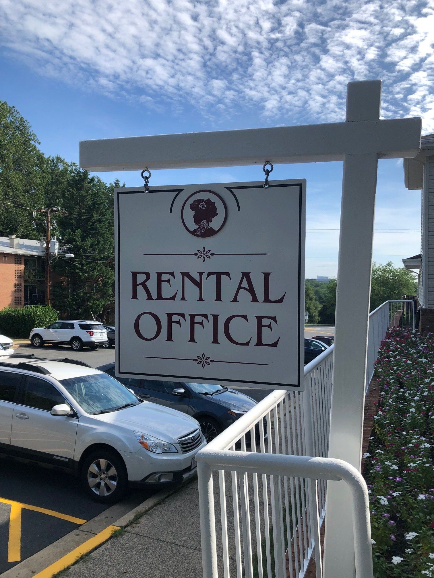 A rental office sign is hanging from a white pole in a parking lot.