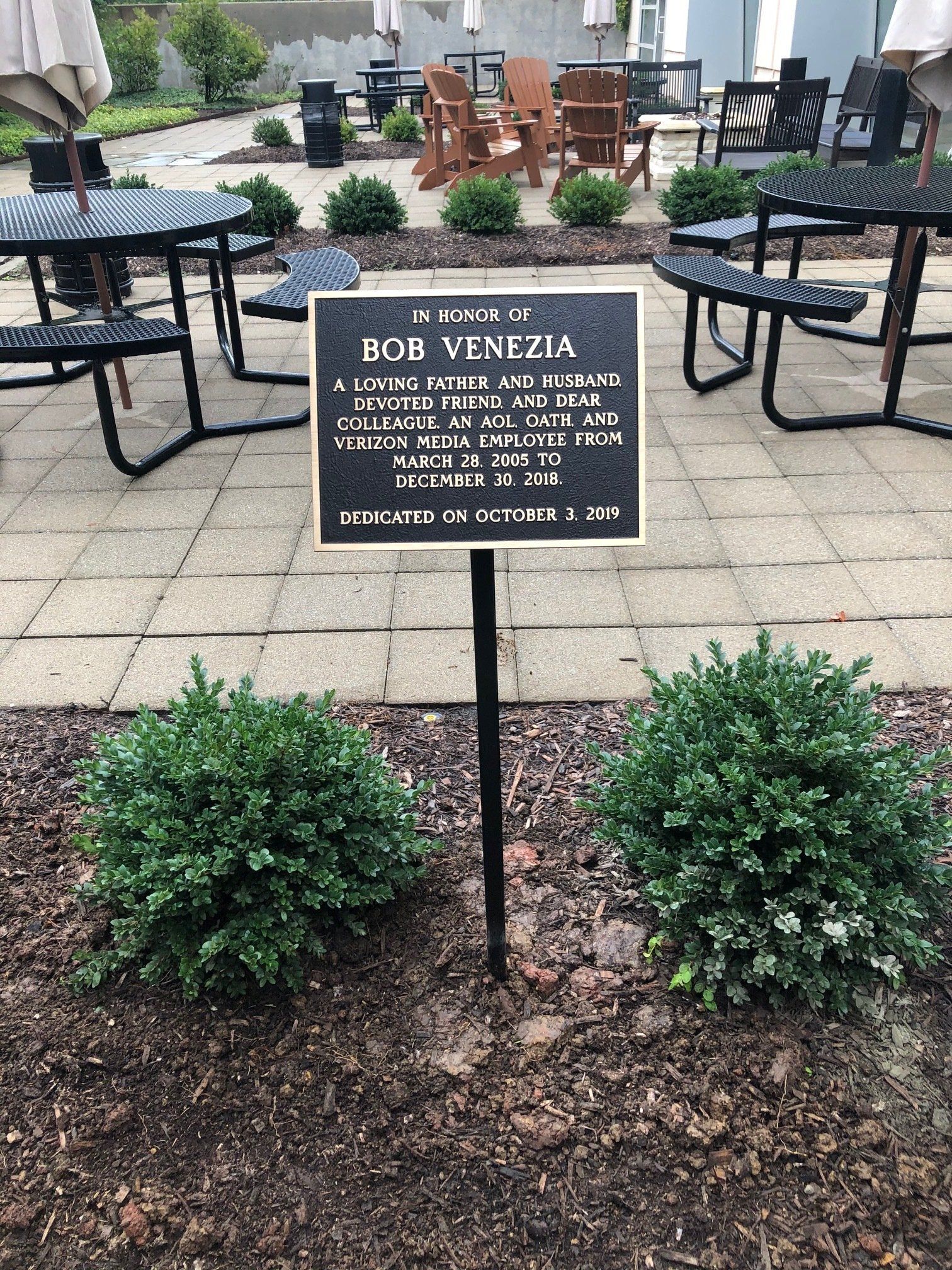 A sign on a pole in front of a patio with tables and chairs.