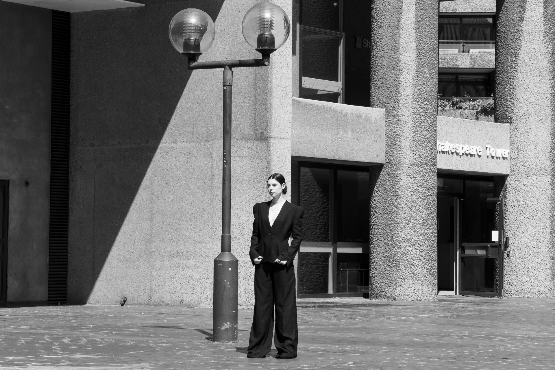 Woman in a suit stands near a street lamp in front of a Brutalist building.