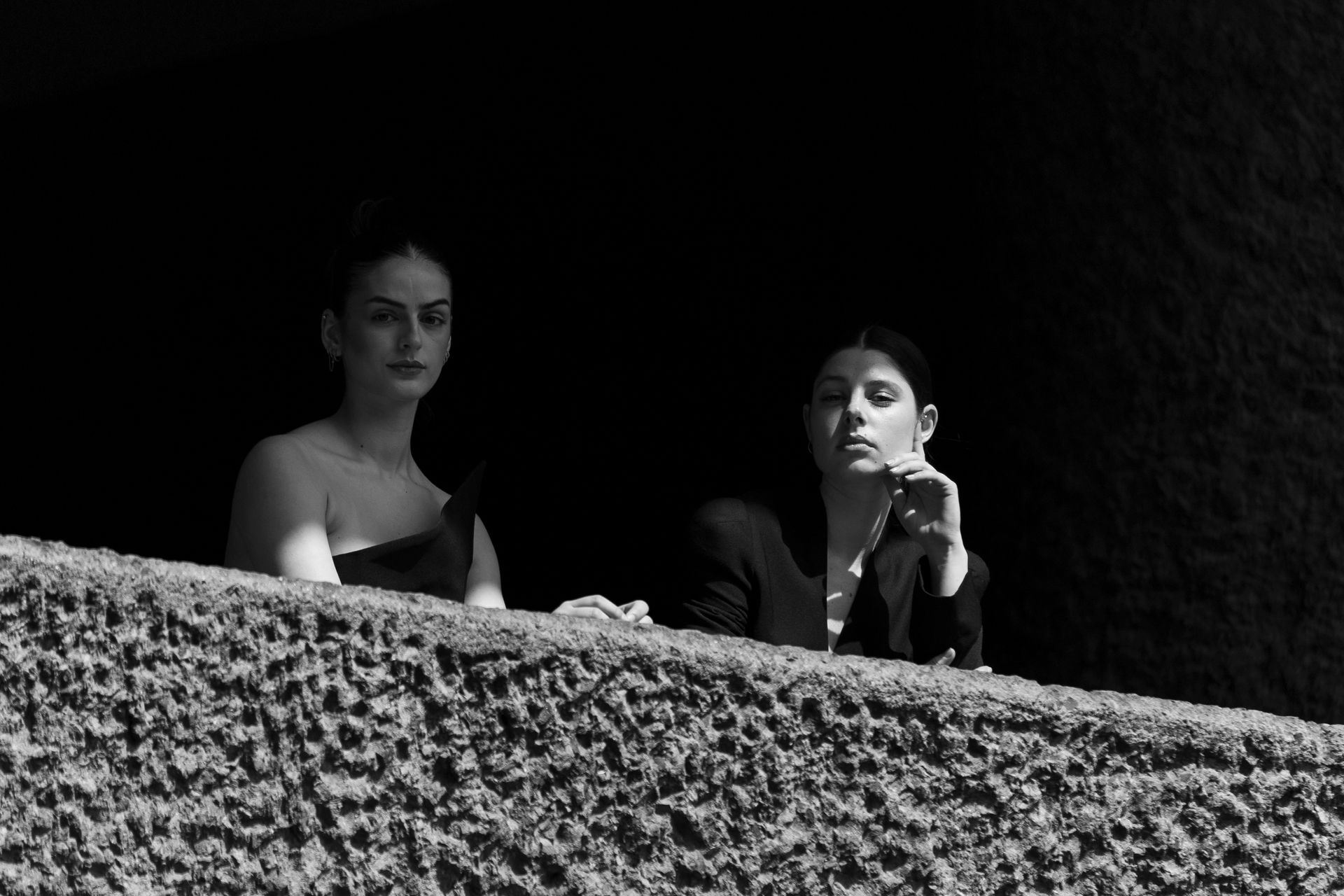 Two women stand behind a textured wall. One in an off-shoulder dress, the other in a blazer, both look toward the camera.