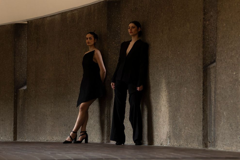 Two women in black outfits lean against a concrete wall. One in a dress, one in a suit.