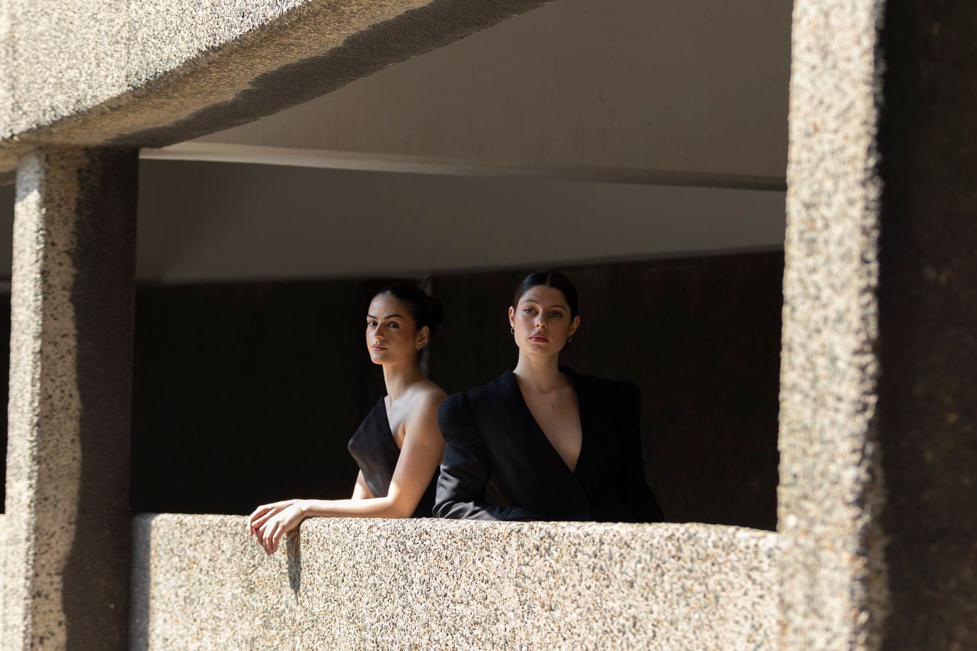Two women in dark clothing stand on a concrete balcony, one facing the viewer.