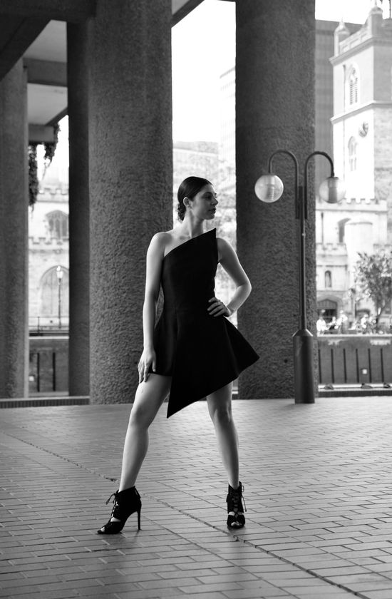 Woman in a black asymmetrical dress and heels poses outdoors, hand on hip.