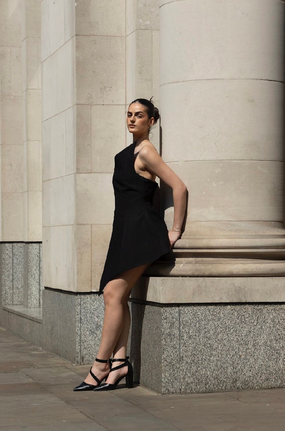Woman in black dress leans against a stone column, wearing sandals; outdoors.