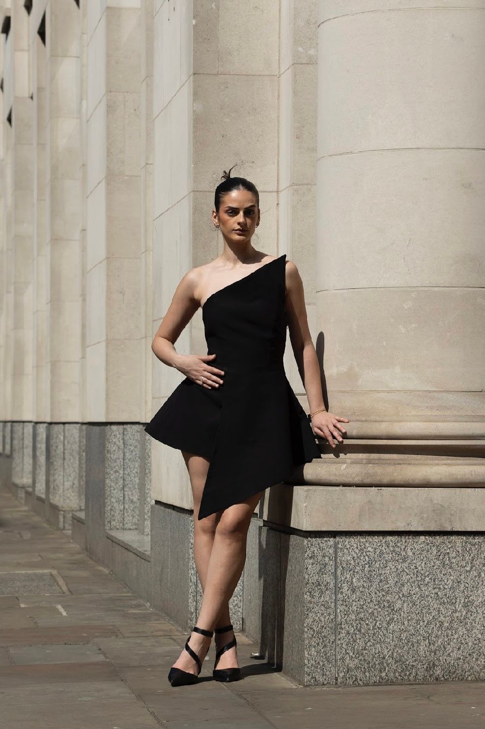 Woman in black asymmetrical dress and heels leans against pillar outdoors.