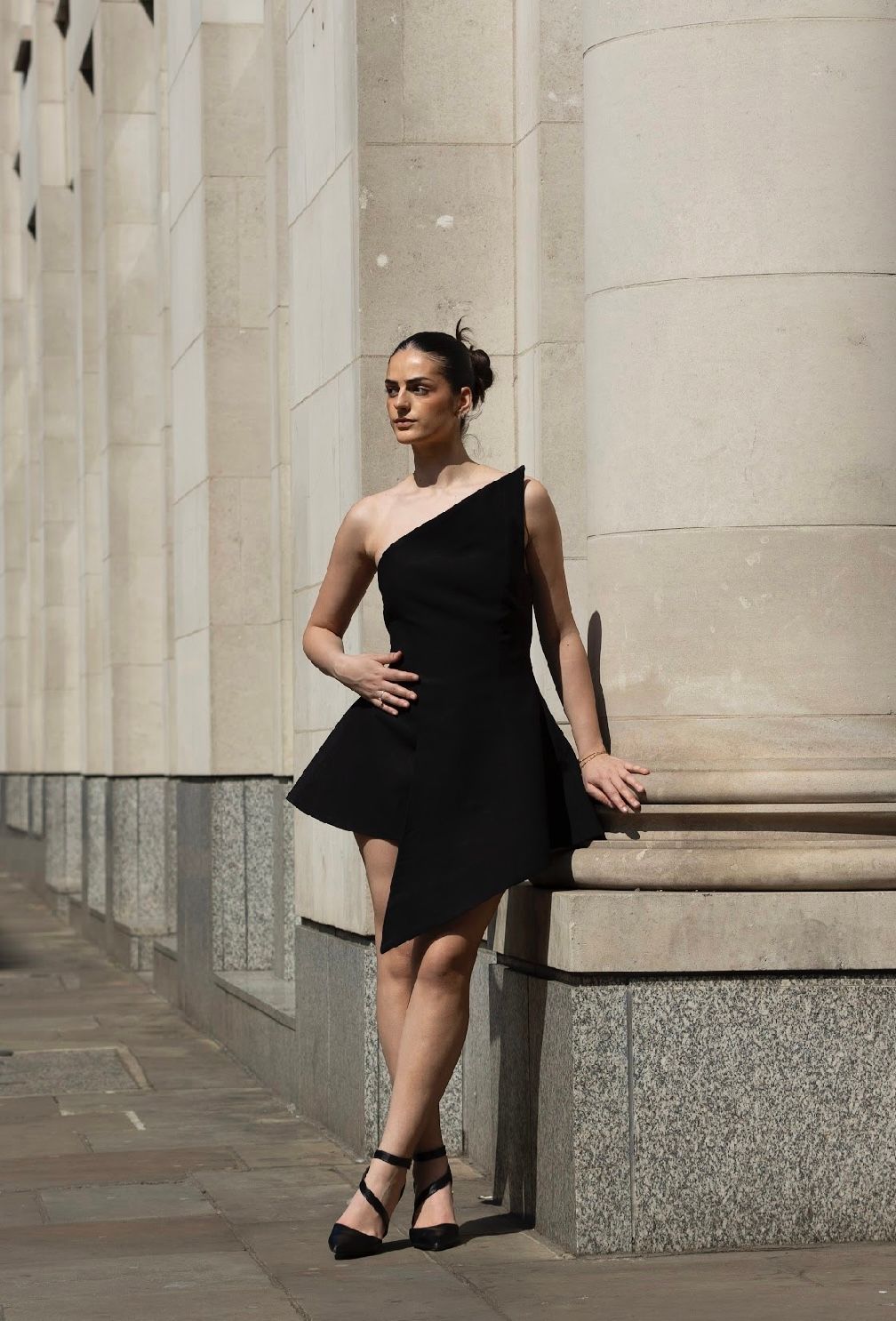 Woman in black one-shoulder dress poses by a column. Outdoors, sunlight.