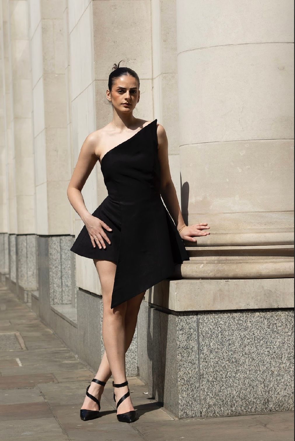 Woman in black one-shoulder dress poses beside a column. She wears heels; sunny outdoor setting.