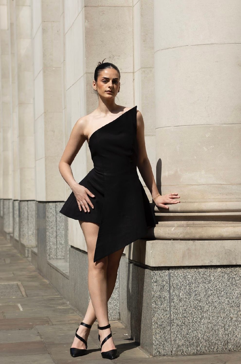 Woman in black one-shoulder dress poses beside a column. She wears heels; sunny outdoor setting.