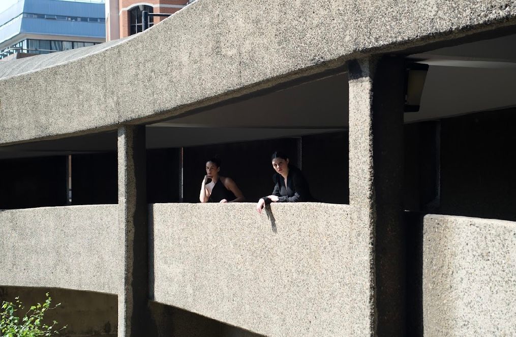 Two women leaning on a concrete ledge, looking out. Sunlight, modern architecture.
