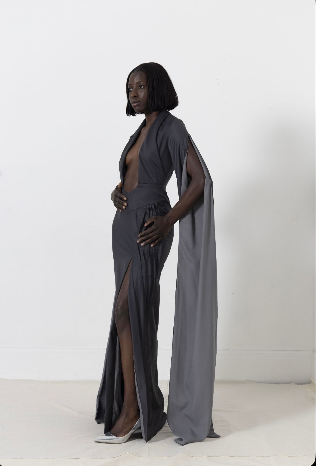 Woman in a dark gray gown with long sleeves and a plunging neckline, stands against a white background.